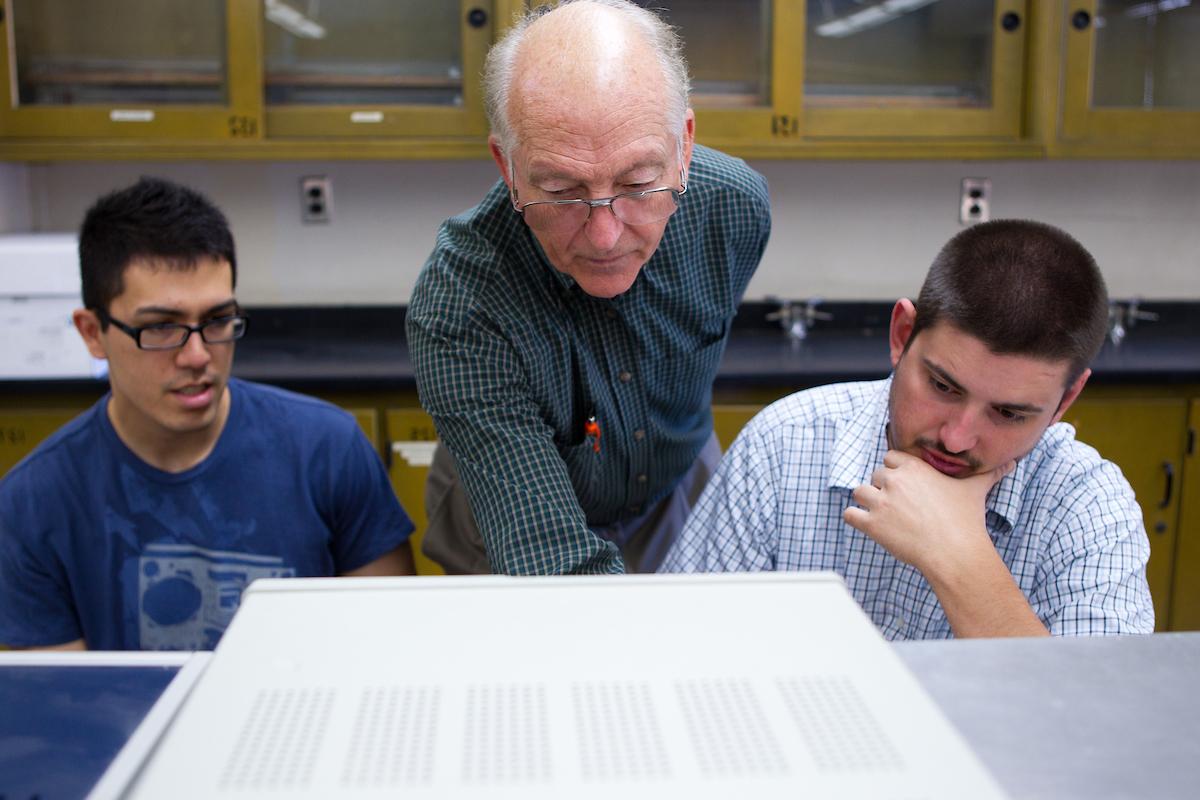 A professor stands between two students and helps them with an assignment.
