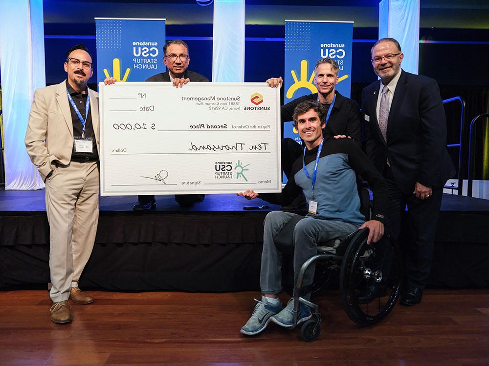 A male, college-age startup founder in a wheelchair poses with two sponsors, two hosts, and a giant check for $25,000