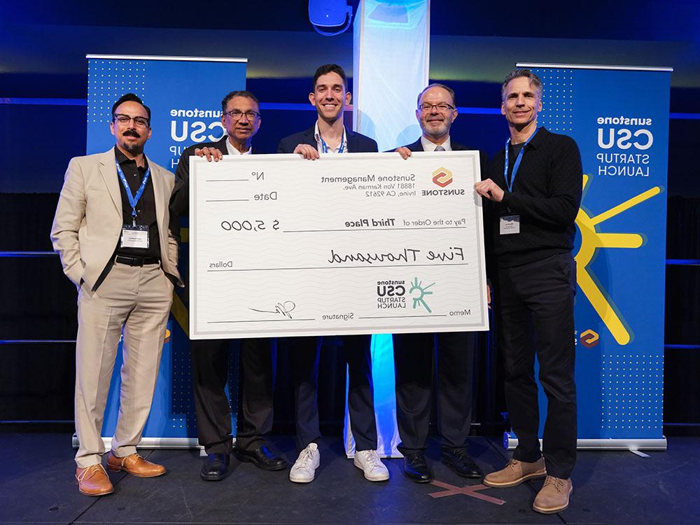 A male, college-age startup founder poses onstage with two sponsors, two hosts, and a giant check for $25,000